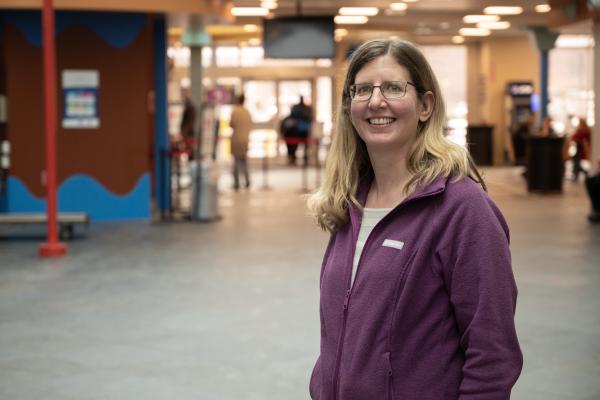 Photo of Mary Ellen Rainey. A woman stands smiling at the camera. She has long blonde hair, glasses and she's wearing a purple jacket.