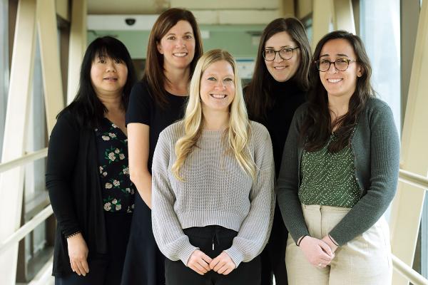 A group of five women stand together in an enclosed walkway with large windows. They are dressed in business casual attire. The woman in the center has long blonde hair and is wearing a light gray sweater. The others are dressed in various dark and neutral tones, with different hairstyles and glasses.